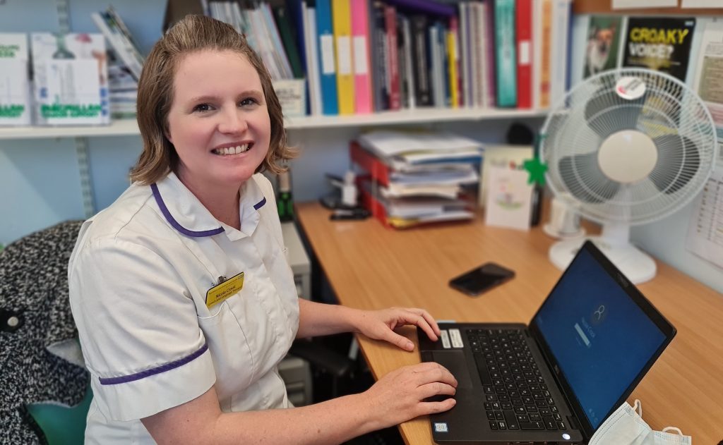 Specialist Speech and Language Therapist (SLT) Dr Nicola Crook seated at a desk in her clinic. In the background are book shelves and a laptop is on the desk in front of her.