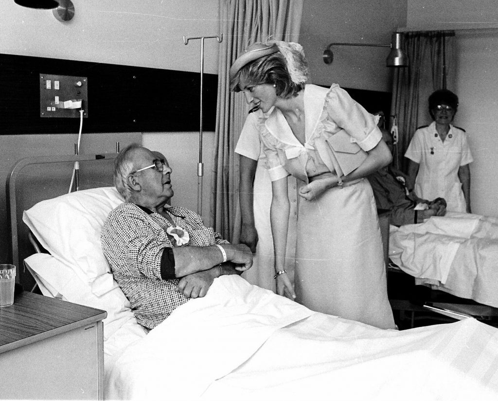 Princess Diana is pictured on a ward, speaking to a patient who is laying in bed. Behind her is another patient and a nurse