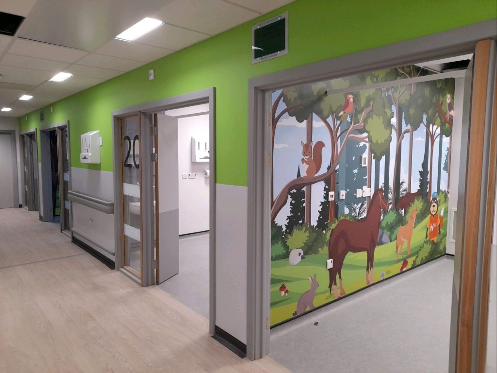 A corridor with four paediatric cubicles opening off it. Through the open door of the first room, we can see a forest mural with trees and animals