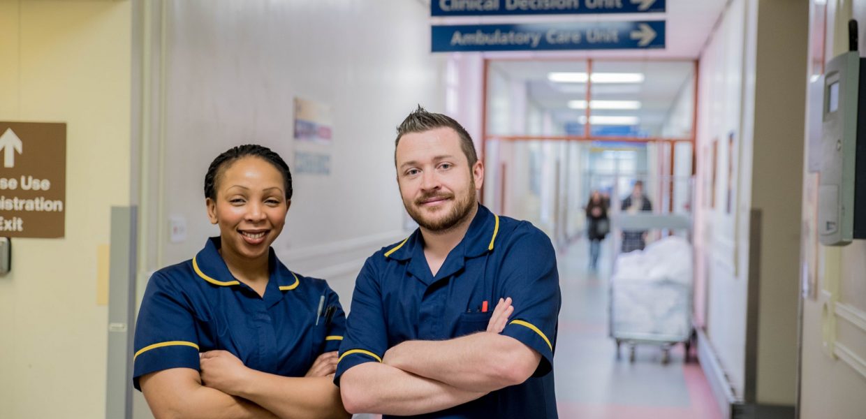 Two clinicians wearing dark blue tunics with yellow detailing, pictured in a hospital corridor. A trolley containing laundry can be seen in the background.