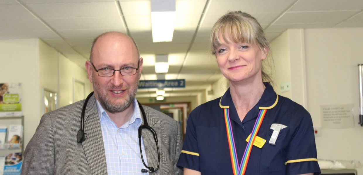 A man with a stethoscope around his neck is standing next to a female nurse. The picture was taken before the COVID-19 pandemic which is why they are not social distancing or wearing masks.