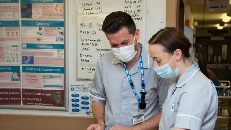 Two healthcare assistants stand at a nursing station looking down at paperwork