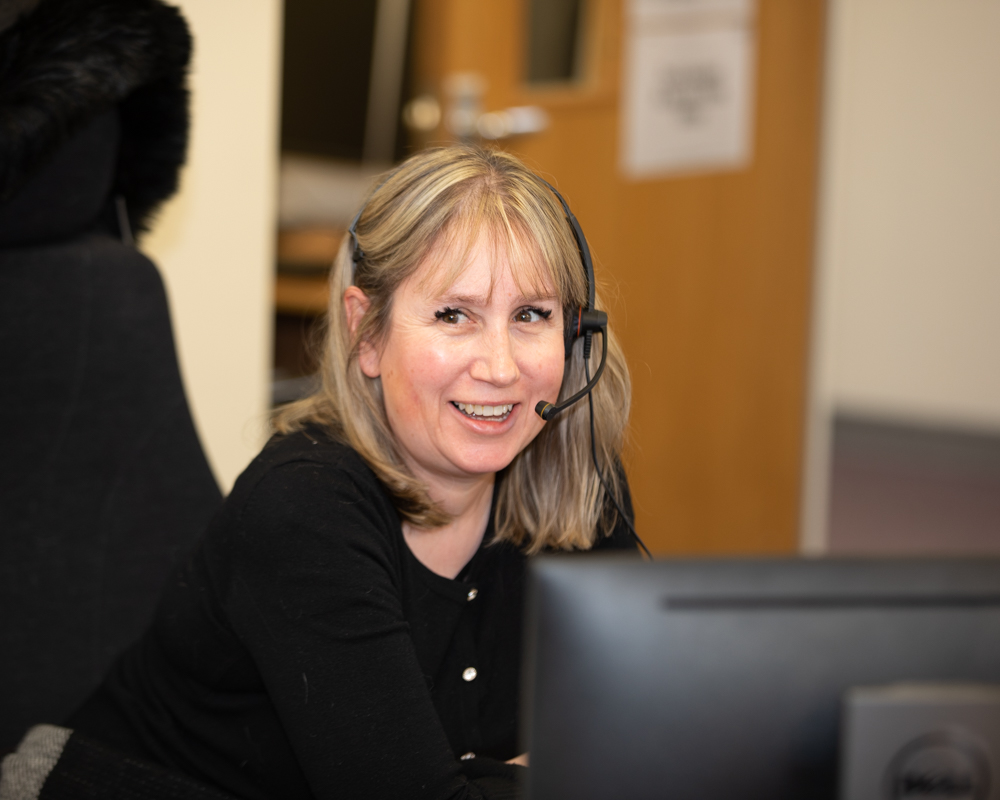 A woman with a headset, sitting at a desk and smiling