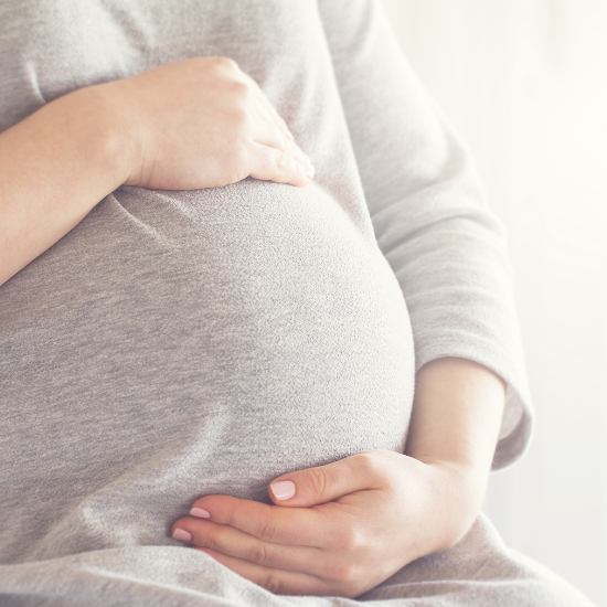 A close up of a pregnant woman's belly. She is wearing a grey long-sleeved top and her hands and encircling her bump