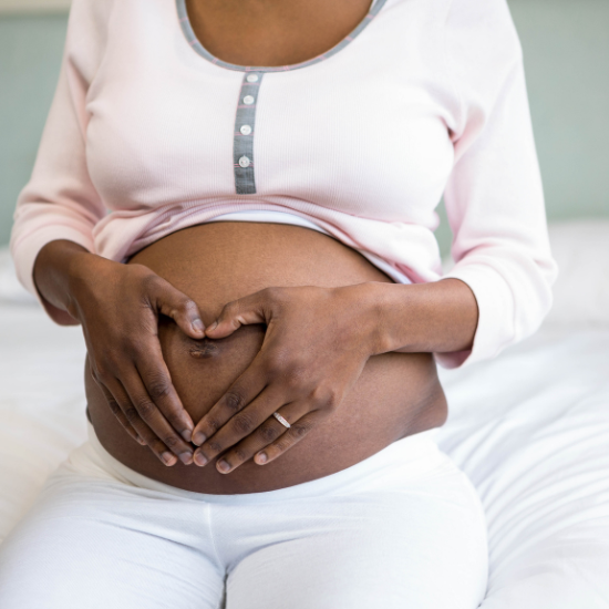 A close up of a woman's pregnant belly. She has pulled up her top to show her bump and her hands have formed a heart shape around her belly button