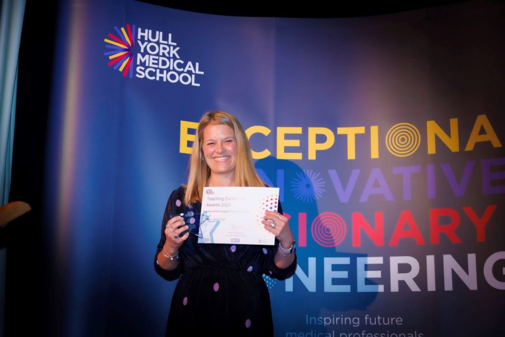 A smiling woman is holding up a certificate. The picture is of Heidi Wright who won the Admin Support worker award at the Hull York Medical School awards ceremony. 