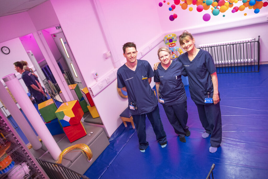 Three physiotherapy staff standing in the new sensory round surrounded by colourful toys and pink lighting