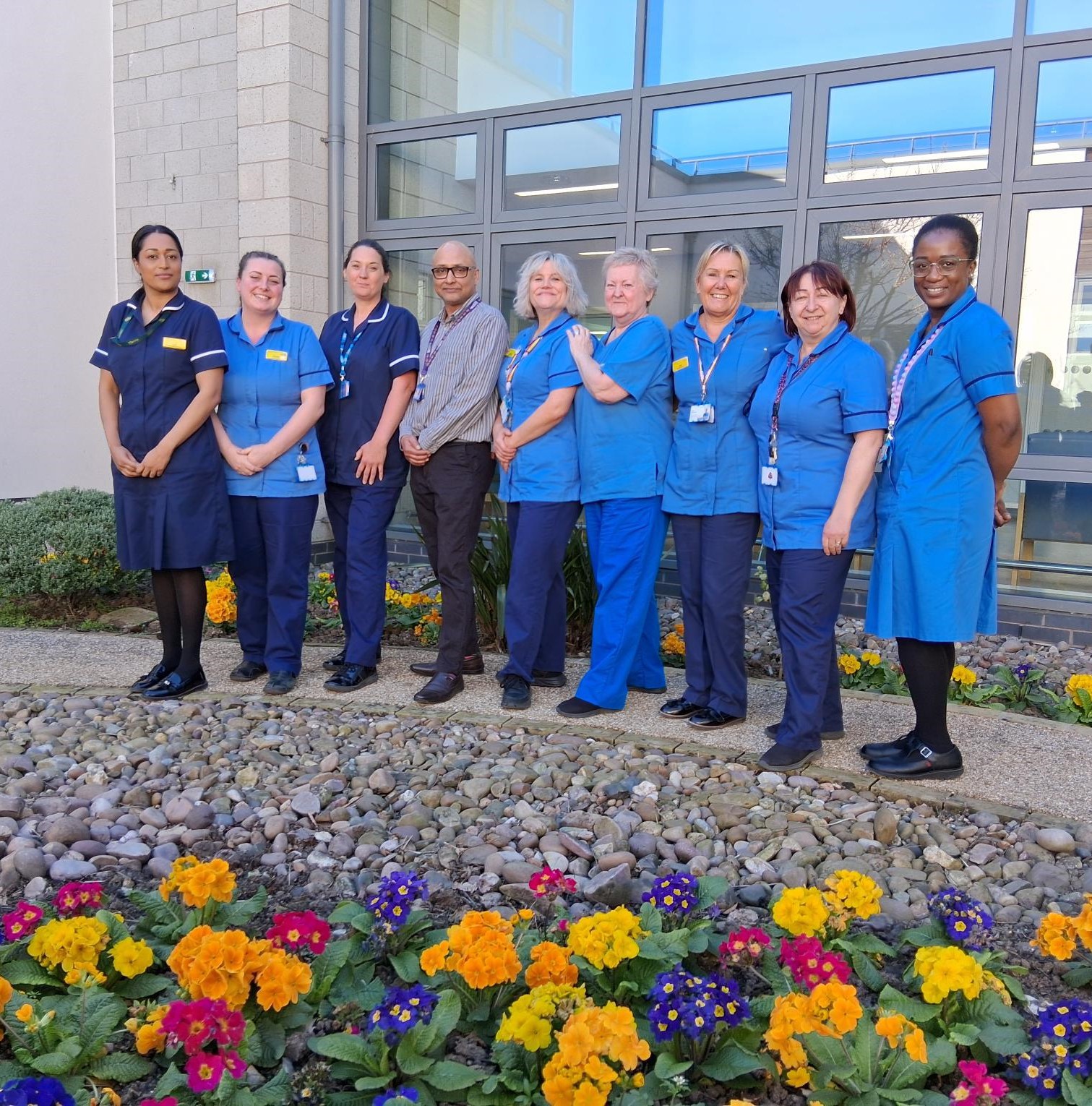 A GROUP OF NURSES STANDING OUTDOORS IN BLUE UNIFORM. tHERE ARE FLOWERS IN THE FOREGROUND OF THE PICTURE.