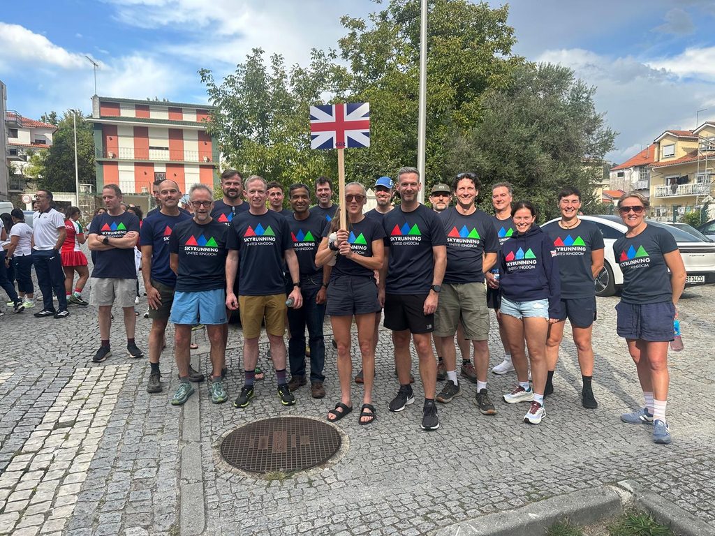 Skyrunning Masters Team UK pose in a Portuguese square. They're wearing matching navy tops with stylised mountains on the front and holding a union flag on a pole.