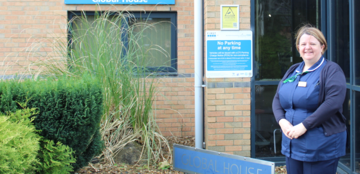 A woman in a nurses blue uniform is standing outside the front of a building which has a sign in the background saying 'Global House'.