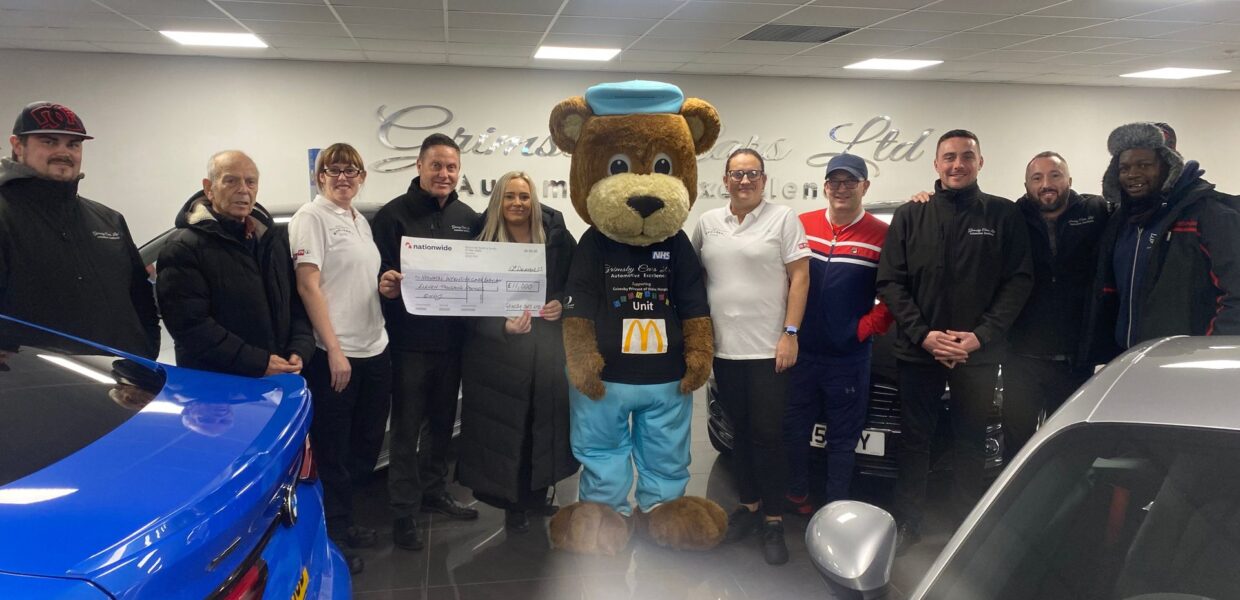 Men and women in a car showroom for a cheque presentation. In the middle, is Scrubs the Bear