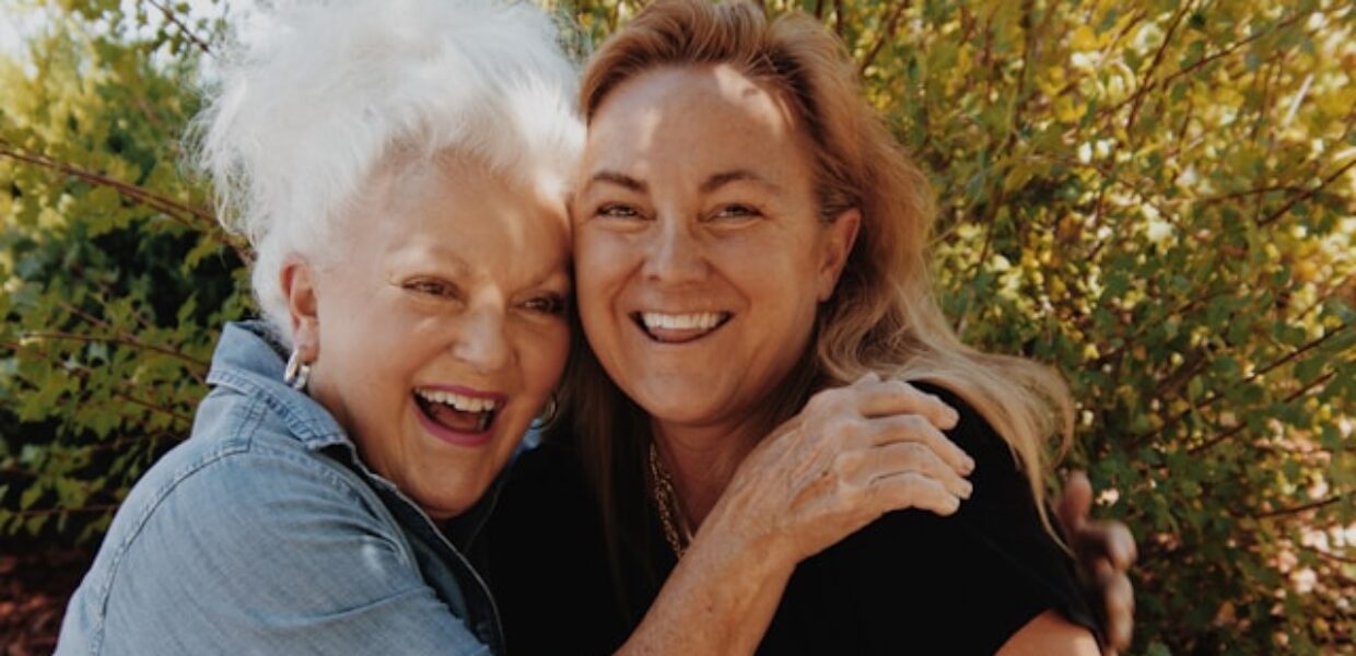 Two women stand in front of a tree, hugging each other and smiling