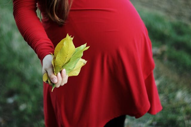 A pregnant woman with long hair and wearing a red top holds autumn leaves in her hand