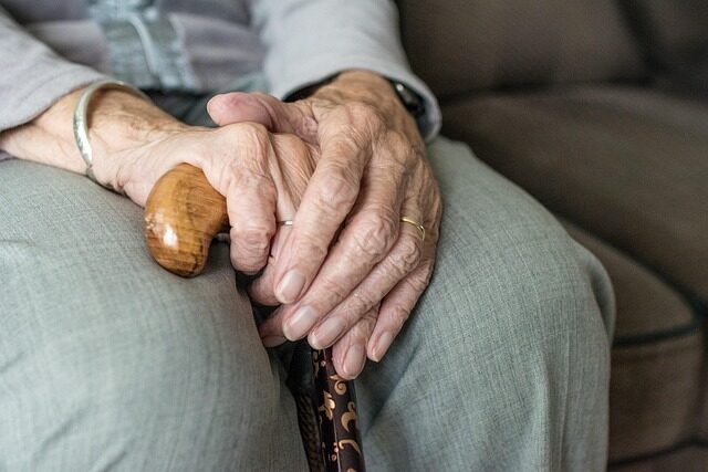 Older person sits on a chair, holding a walking stick