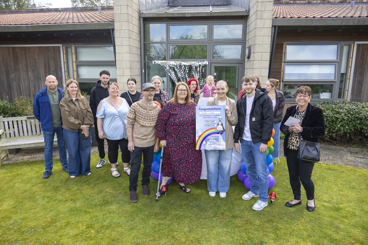 Lilli Hales holds the end of treatment bell in a garden surrounded by family and friends.