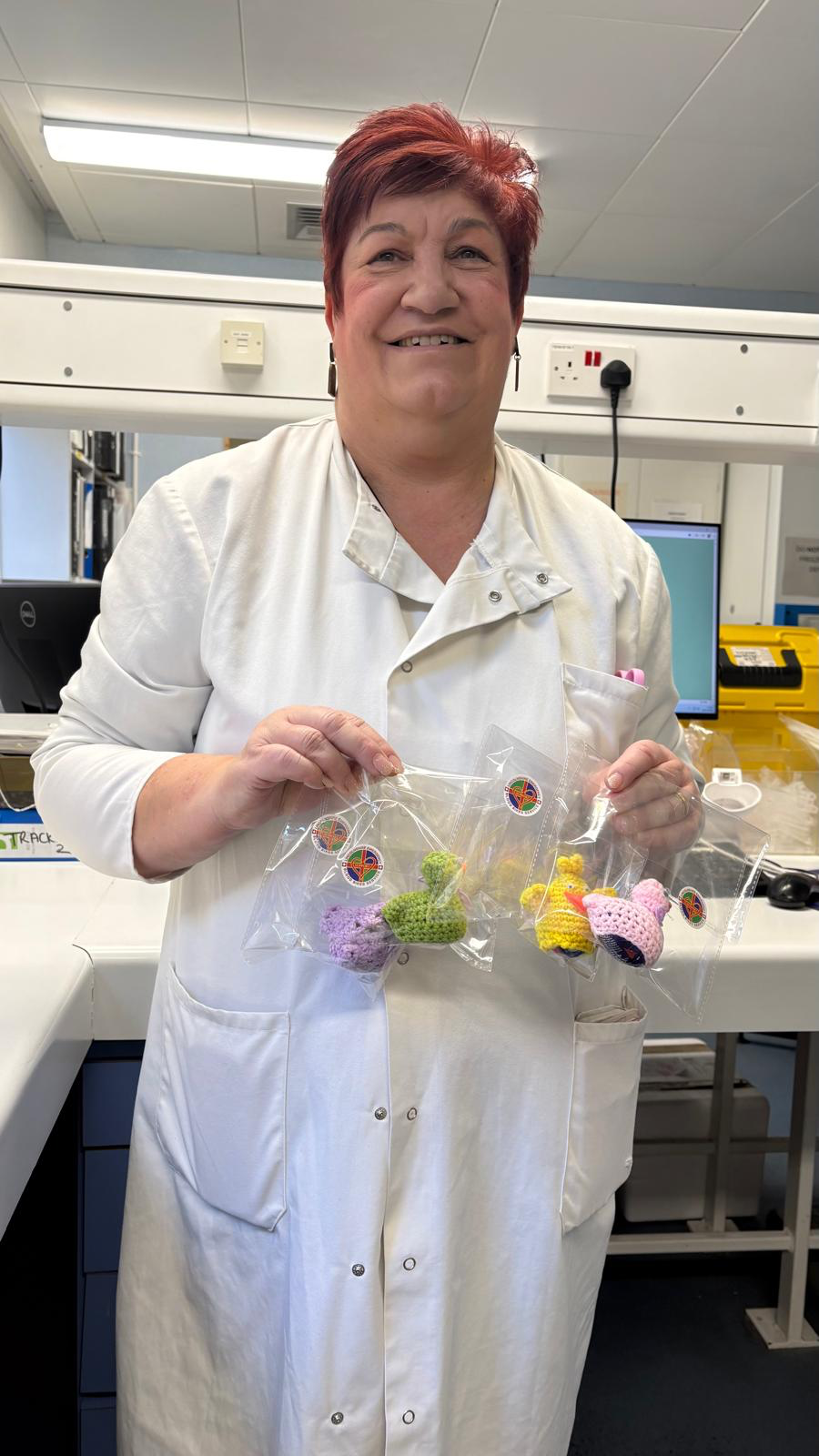 Glenda poses in her white lab coat, holding the crocheted items she has made to raise money for the Lincolnshire Emergency Blood Bikes