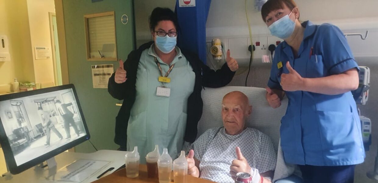 Two nursing staff stood next to a patient in bed with their thumbs up