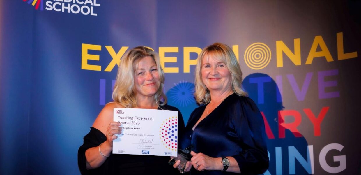 Two women smiling proudly holding a certificate from the Hull York Medical School awards ceremony. In the background is the HYMS logo.