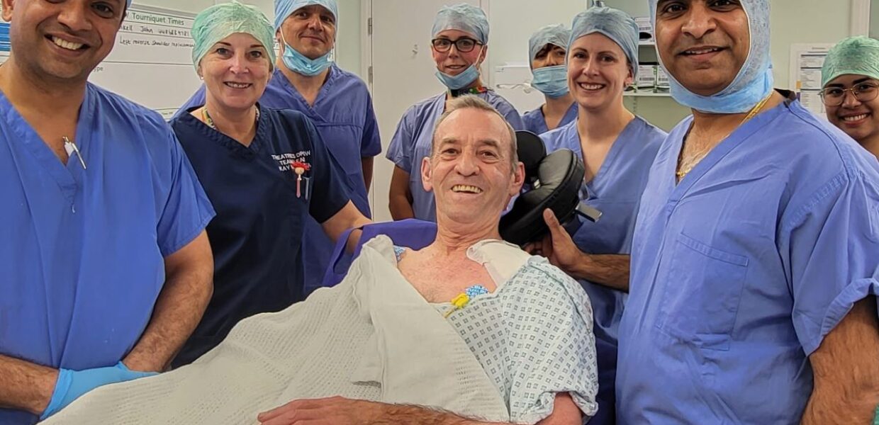 A man in a hospital theatre gown is lying on a trolley smiling. He is surrounded by eight other people - men and women - who are dressed in blue theatre scrubs