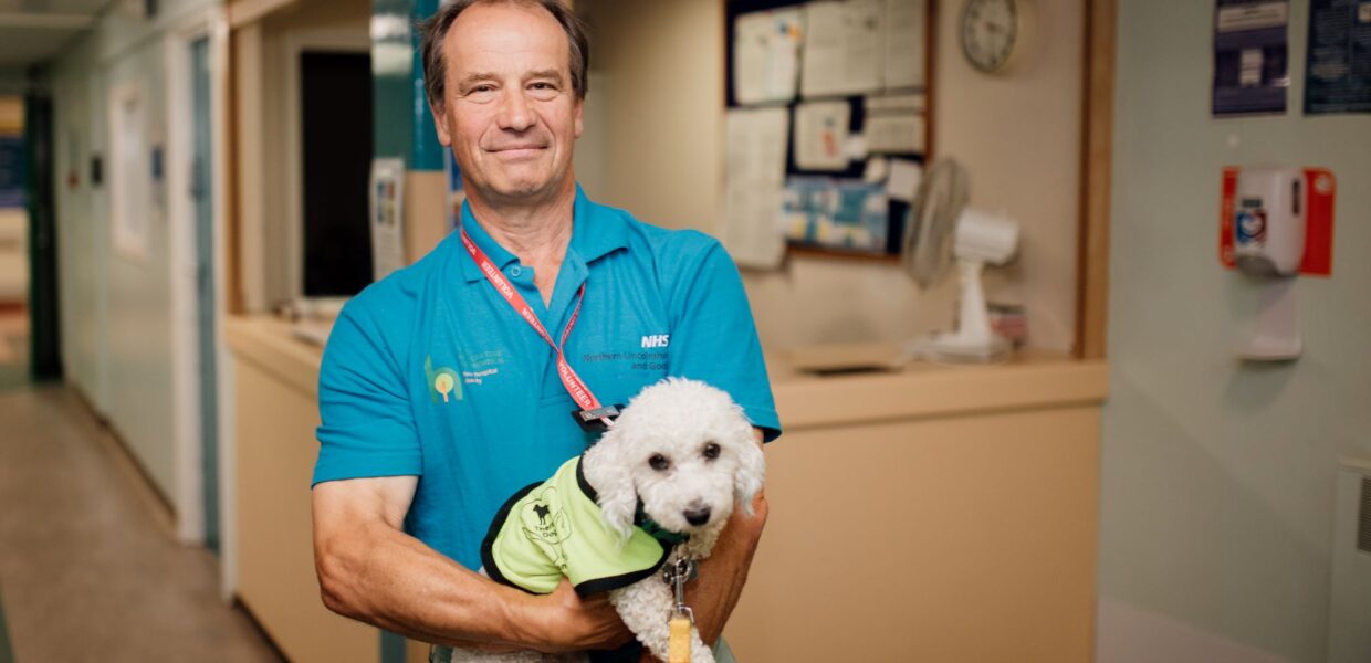 a volunteer in a blue top smiles and holds a therapy dog