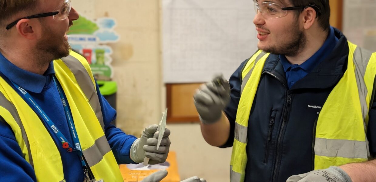 Maintenance Assistant Josh and student, Regan, laugh together in the Estates workshop. Both are wearing blue jumpers, high vis vests, safety glasses and gloves. Regan has a piece of wood in a vice in front of him, which he has been marking up and cutting down to Josh's instructions