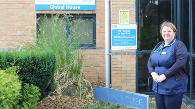 A woman in a nurses blue uniform is standing outside the front of a building which has a sign in the background saying 'Global House'.