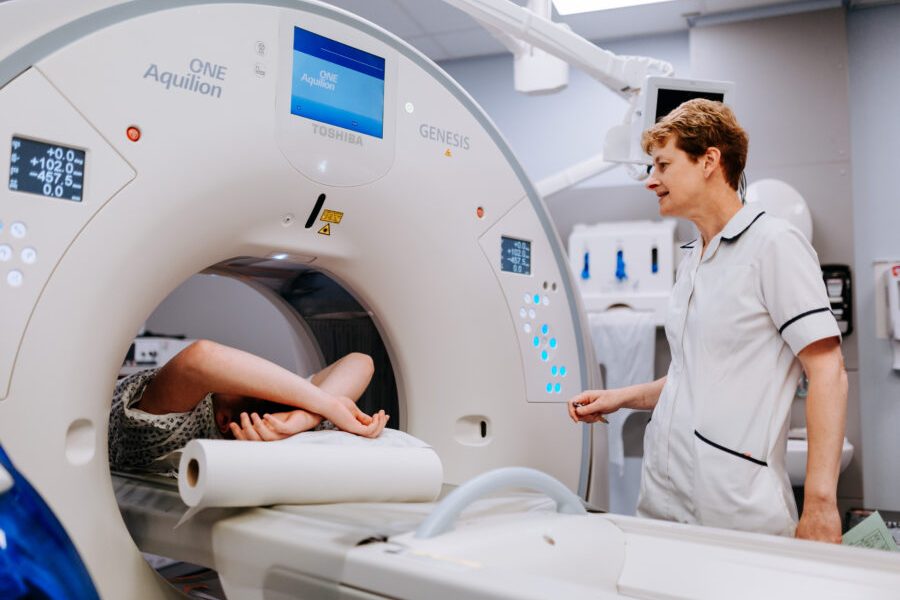 a patient laid down with their arms above their head inside a scanner with a member of staff watching on