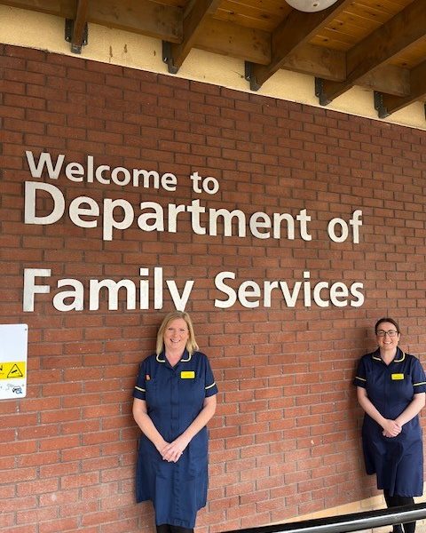 two midwives in blue uniform stood next to a brick wall with a sign reading 'welcome to the department of family services
