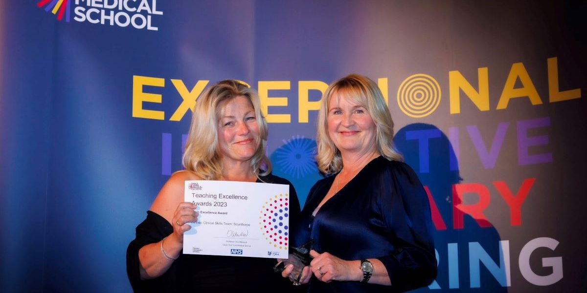 Two women smiling proudly holding a certificate from the Hull York Medical School awards ceremony. In the background is the HYMS logo.