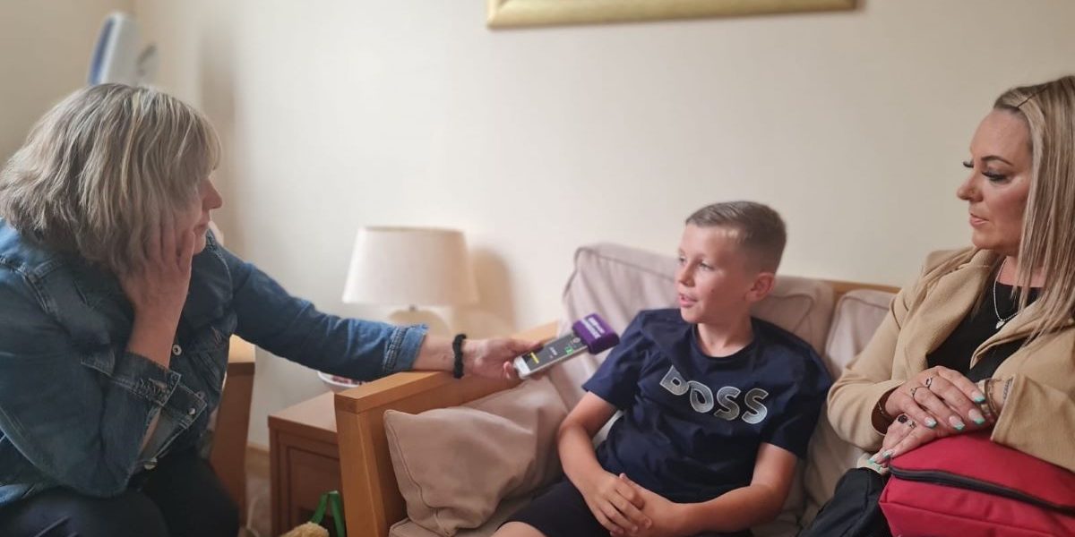 There is a woman sitting down holding out a microphone to a young boy sitting on a sofa. Another woman (his mum) is sitting next to him.