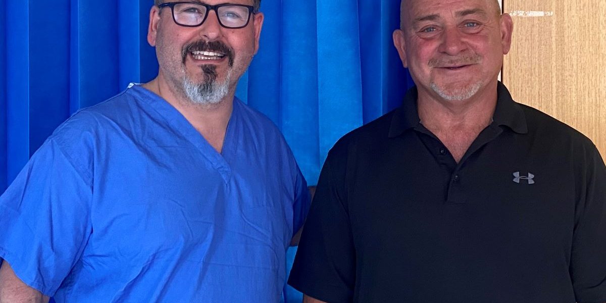 Two men, one dressed in blue theatre scrubs uniform, are standing next to each other