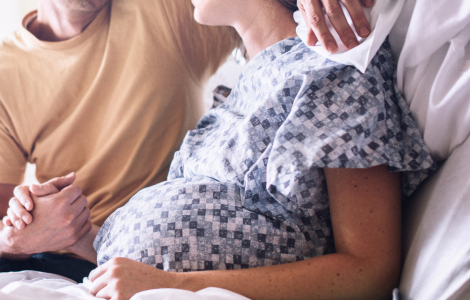 A pregnant woman sits up in bed, resting on pillows. A man has his arm around her.