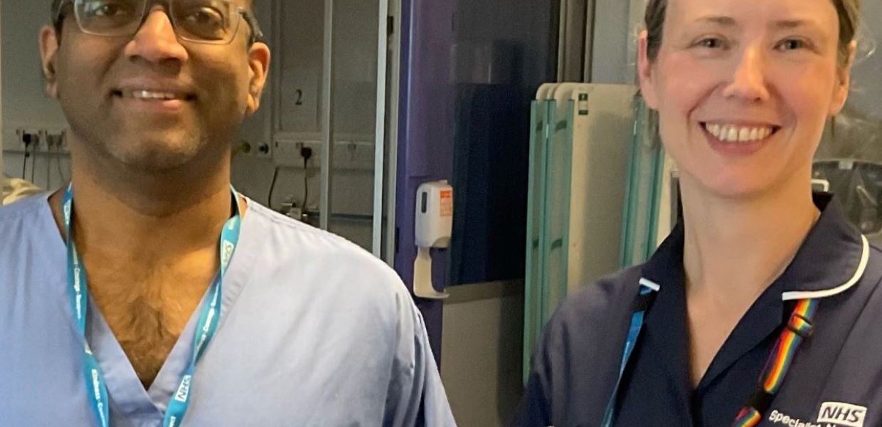A man and a woman standing side by side smiling. They are in a clinical area and are wearing clinical blue uniforms.