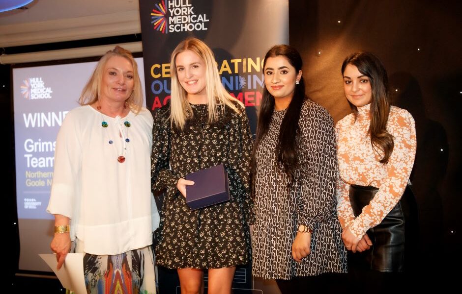 Four women pictured at an awards ceremony in front of a HYMS pull-up banner.