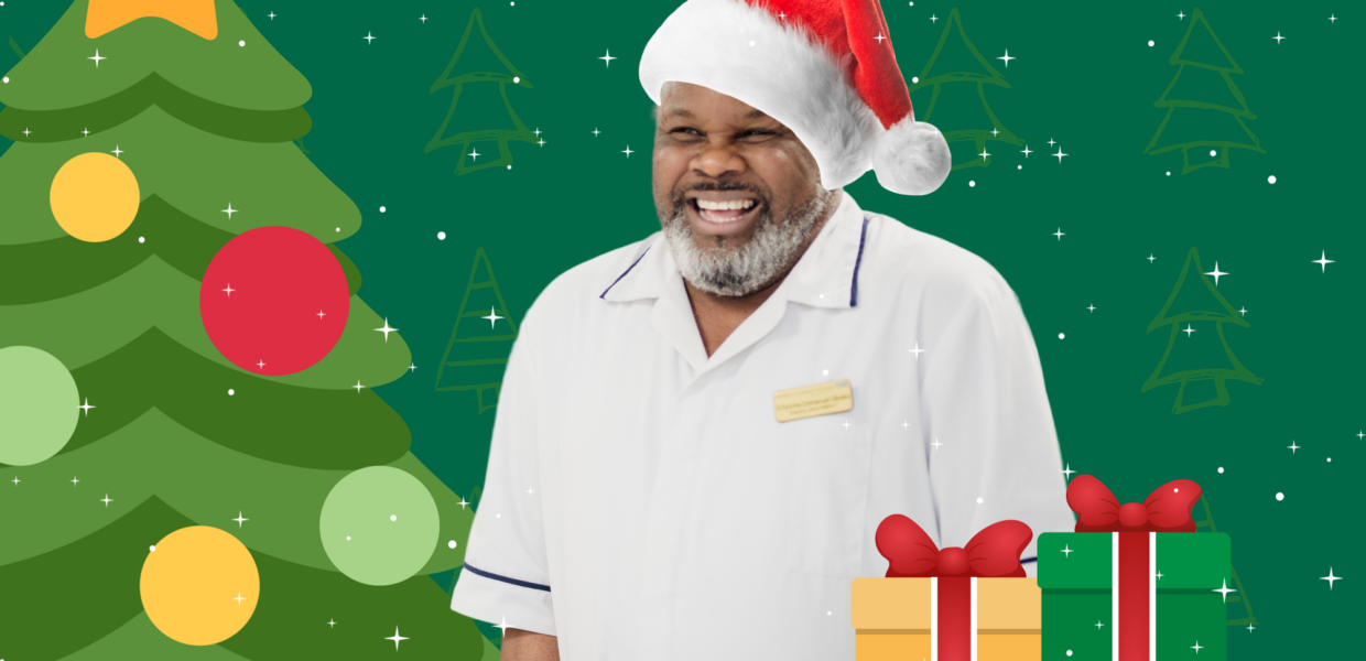 A member of our staff, smiling broadly, and wearing a santa hat. In the background is a Christmas tree and to the side of him, a pile of wrapped presents