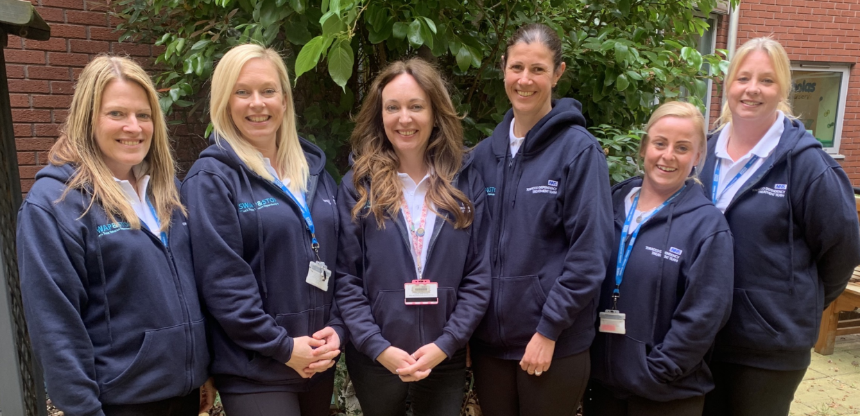 Members of the Tobacco Dependency Treatment Service team pose in matching navy jackets in front of a green leafed bush and red brick wall